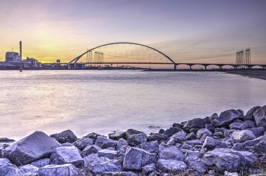 Yeni köprü De Oversteek (Crossing) Nijmegen, Hollanda bir kayalık sahil ön planda ile doğru nehir Waal görünümünü akşam