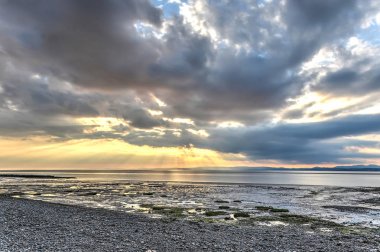The rays of the late afternoon sun break through the clouds over the waters and adjacent mudflats of the bay of Morecambe, Lancashire, England clipart