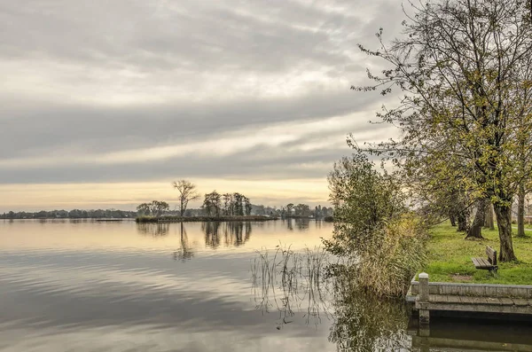 Tranquil scene with a small bench overlooking one of the Reeuwijke Plassen (Reeuwijk Lakes) under a dramatic cloudy sky on a calm day in november
