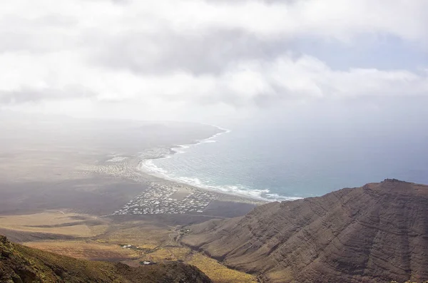 North Coast Of The Isla De Lanzarote, Spain, View Over The Bluff Of The Famara Massif With The Village Of The Same Name Down At The Coast Line.