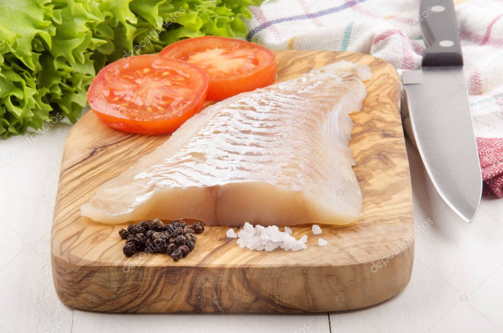 raw haddock fillet with tomato slices and pepper on wooden cutting board  