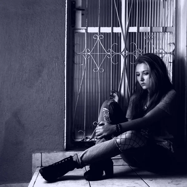 Black and white image of a long hair sad girl seated at the doorstep of her home.