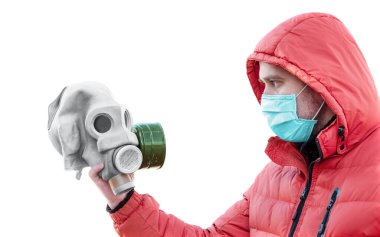 Tıbbi maskeli ve kapüşonlu bir adam elinde beyaz bir arka planda gaz maskesi tutuyor.