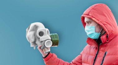 Tıbbi maskeli ve kapüşonlu bir adam elinde mavi bir arka planda gaz maskesi tutuyor.