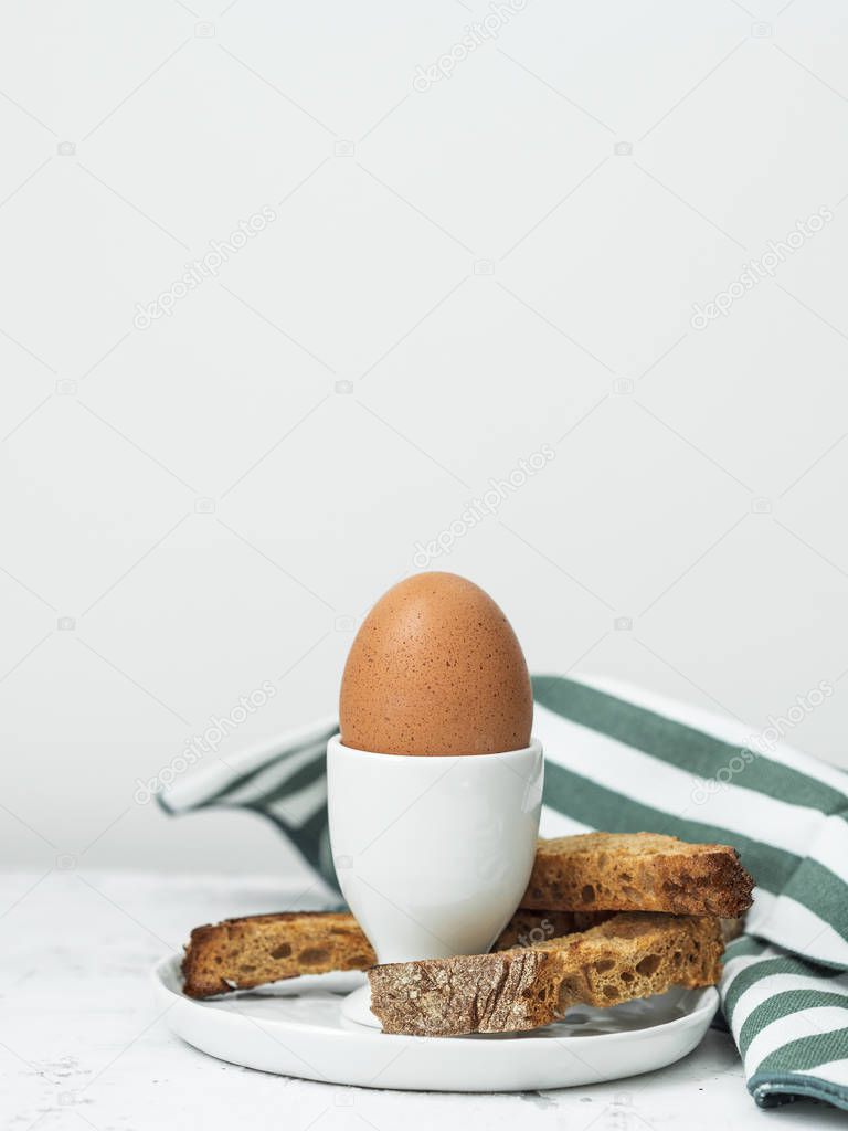Boiled egg in eggcup served with croutons on white stone table with striped towel on background 