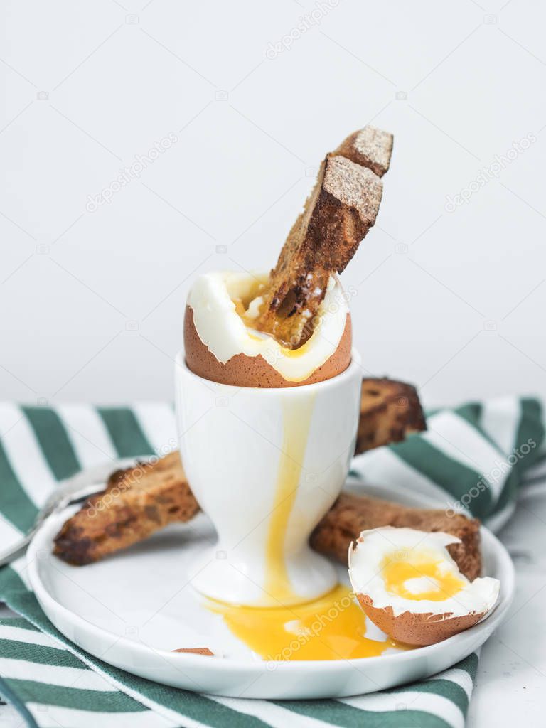 Soft-boiled egg in eggcup with crouton in yolk on white stone table with striped towel on background 
