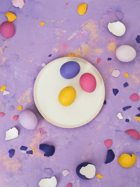 Top view of different colored eggs on white plate and scattered eggshell on purple background