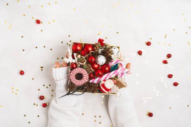 Beyaz örülü süveter giyen kadın elleri Noel oyuncaklarıyla dolu bir kutu tutuyor beyaz arka planda kopyalanmış ağaç oyuncakları. Noel ve Yeni Yıl Yaratıcı Tasarımı. 