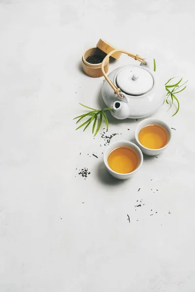 Asya çay konsepti, iki beyaz fincan çay, çaydanlık, çay seti, yemek çubukları, bambu hasırı, beyaz arka planda kuru yeşil çay ile çevrili, yazı için boşluk. Çay demlemek ve içmek..