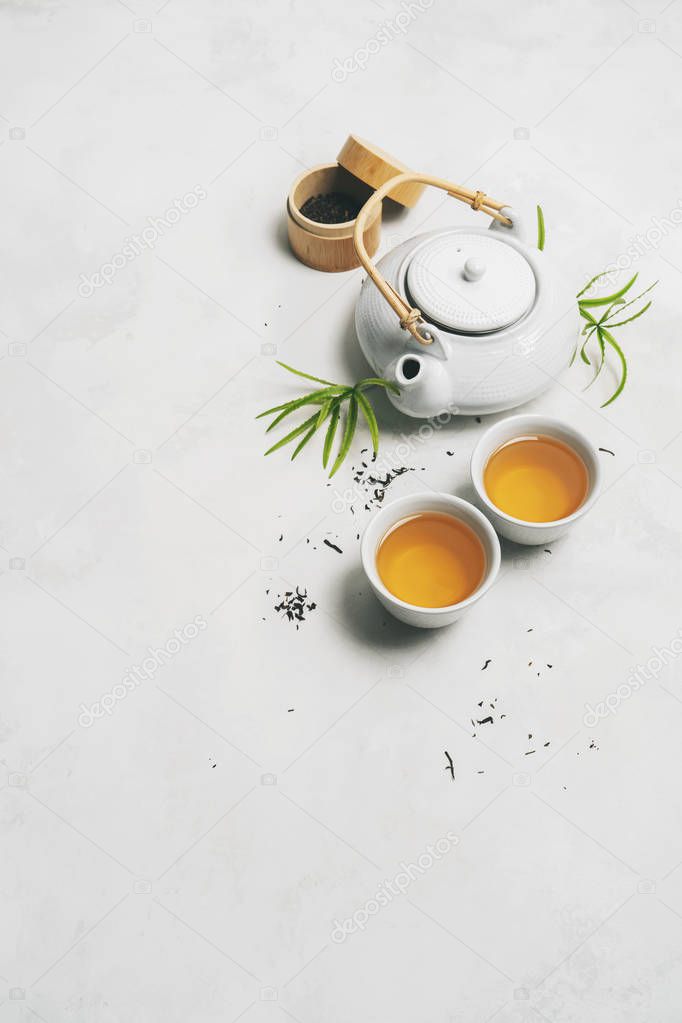 Asian tea concept, two white cups of tea, teapot, Tea set, chopsticks, bamboo mat surrounded with dry green tea on white background with space for text. Brewing and Drinking tea.