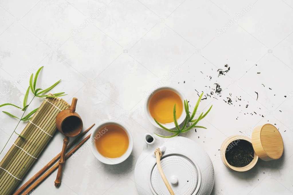 Asian tea concept, two white cups of tea, teapot, Tea set, chopsticks, bamboo mat surrounded with dry green tea on white background with copy space. Brewing and Drinking tea.