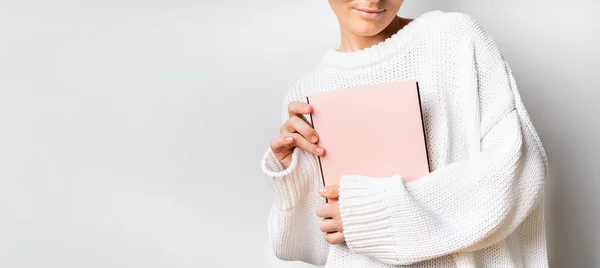 Close view of woman in white woolen sweater holding a book with empty pink cover in hands. Free space for your mock up of reading book concept background.