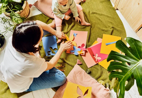 Mom and daughter spend time together imitating summer time on green grass plaid doing papercraft butterflies. What to do at home with children in quarantine.