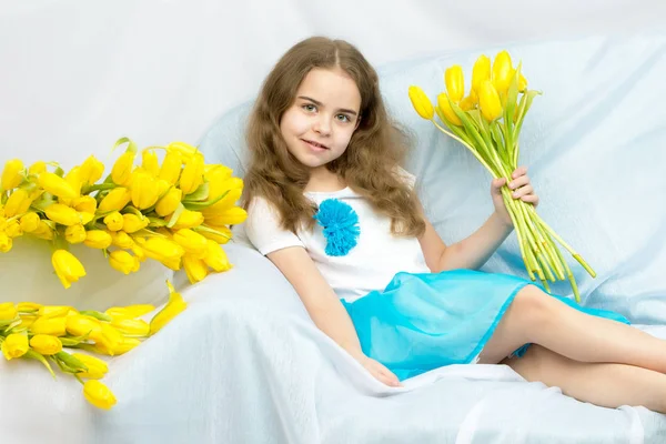 Beautiful blond schoolgirl with yellow tulips sitting on the cou — Stock Photo, Image