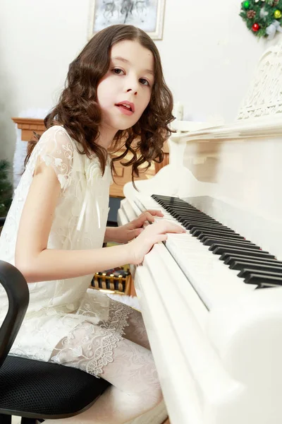 Vacker flicka i en vit klänning som sitter vid pianot. — Stockfoto