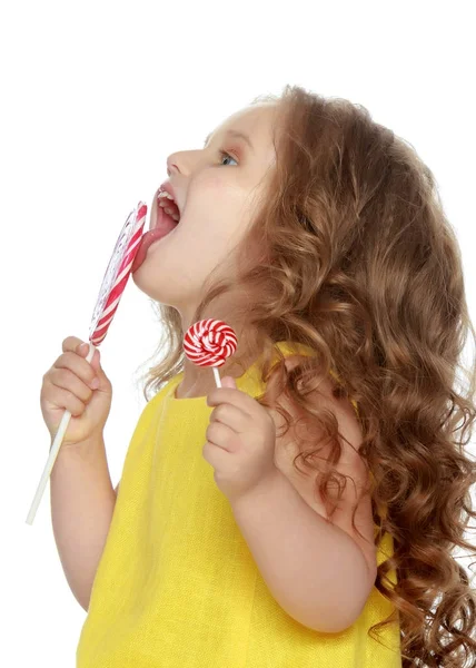 Een klein meisje likt een snoepje op een stick. — Stockfoto