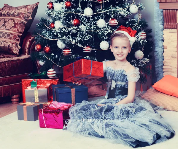 Büyük bir hediye ile Noel ağacının yanında oturan küçük kız. — Stok fotoğraf
