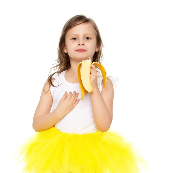 Het kleine meisje in het geel rokje eten van een banaan. — Stockfoto