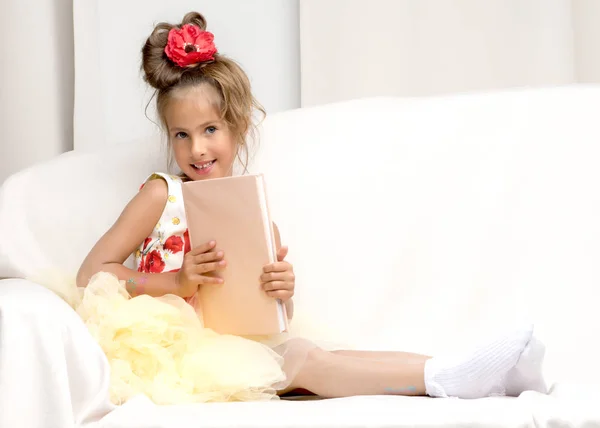 A little girl is sitting on the couch and reading a book. — Stock Photo, Image