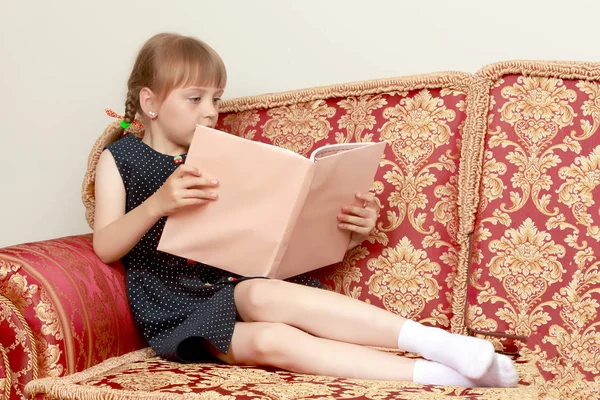 Een klein meisje zit op de bank en leest een boek.. — Stockfoto