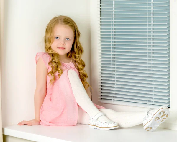 A little girl is sitting on the window on the background of the — Stock Photo, Image