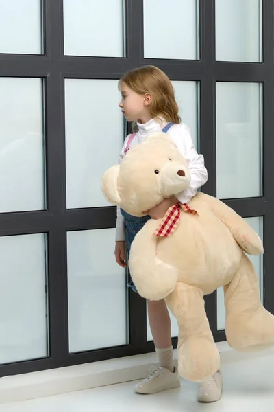 A little girl stands by the window and hugs a teddy bear. — Stock Photo, Image