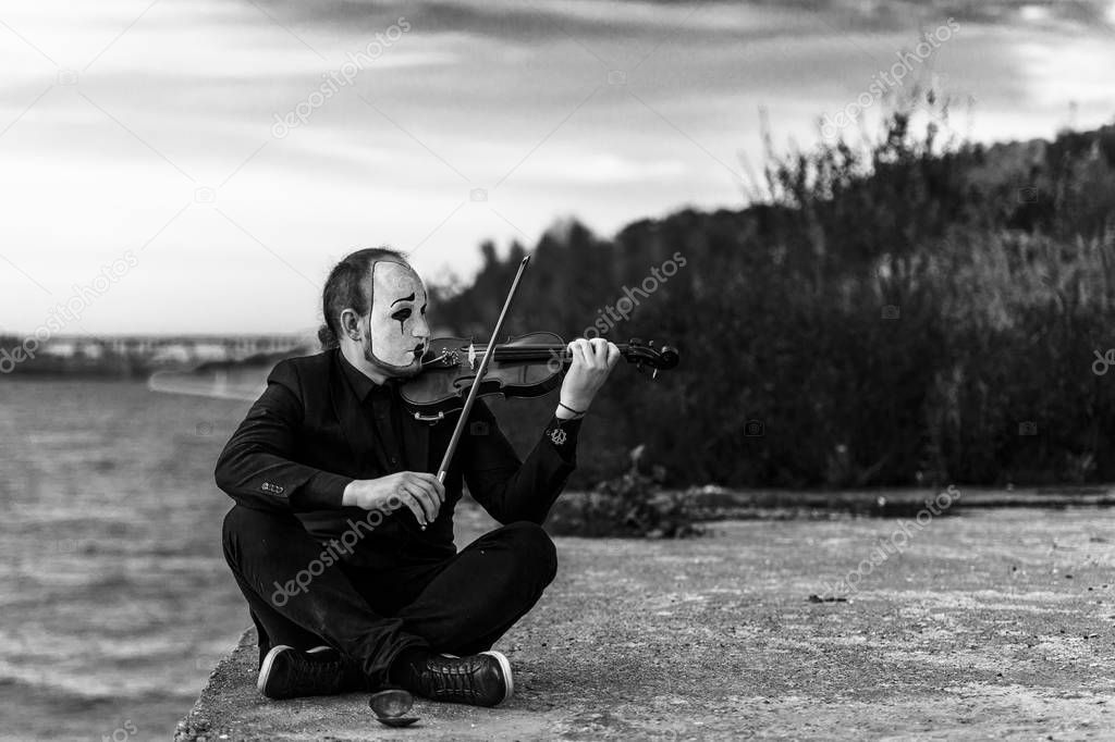 Black and white portrait of mime sitting on riverside and playing violin outdoors