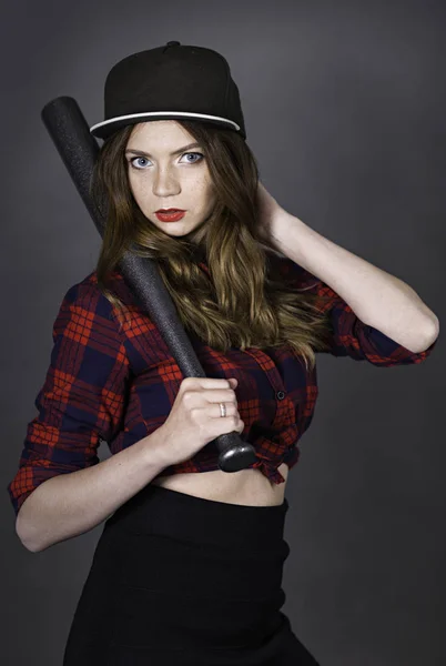 Young attractive woman dressed in baseball cap and red-blue plaid shirt holding pearl-grey bat in hand lay on shoulder