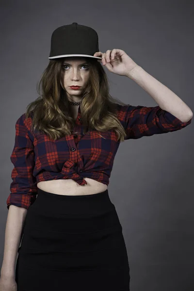Attractive woman dressed in baseball cap, red-blue plaid shirt and black skirt touching cap-peak hand and looking to camera over the grey background