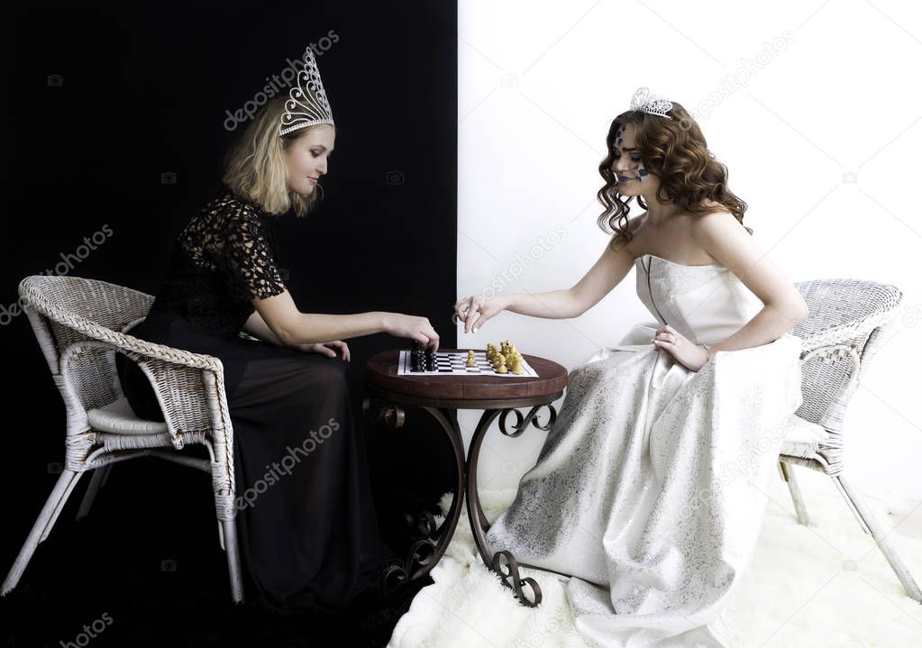 Two girls wearing queen dresses, diamond crown sitting opposite to each other and playing chess.