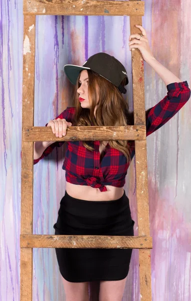 Genç çekici kadın beyzbol şapkası, ahşap merdiven üzerinde dinlenme ve rasgele desen renkli boyalı duvarın üzerinden kameraya bakarak kırmızı-mavi ekose gömlek giymiş
