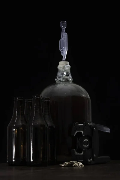 Fermented beer in a demijohn with airlock and brown bottles and over kitchen background — Stock Photo, Image