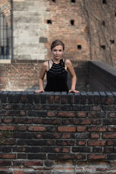 Hermosa chica vestida con vestido negro de pie sobre la pared de ladrillo castillo viejo —  Fotos de Stock