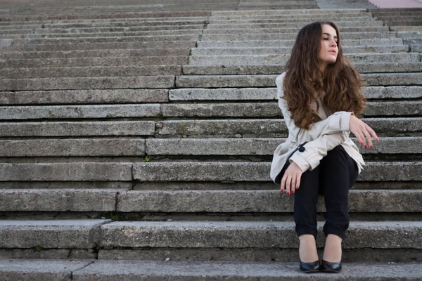 Young beautiful woman wearing beige jacket sitting on concrete stairway and looking away — Stock Photo, Image