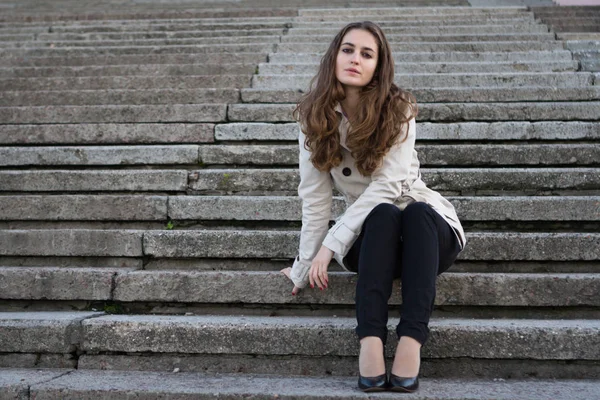 Młoda piękna kobieta noszenia kurtka beżowy, siedząc na beton s — Zdjęcie stockowe