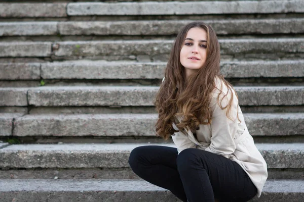 Jovem mulher bonita vestindo jaqueta bege sentado em escadas de concreto — Fotografia de Stock