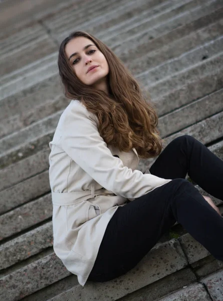Молодая красивая женщина в бежевой куртке сидит на бетонной лестнице — стоковое фото