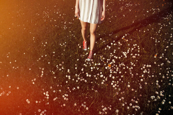 Teenage legs in sneakers and white dress standing on lush springtime grass with flowers, top view.
