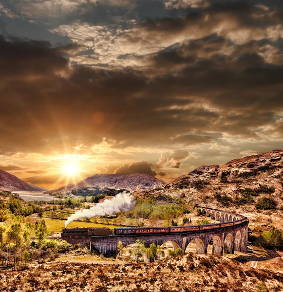 Glenfinnan Viaduto ferroviário na Escócia com o trem a vapor jacobita contra o pôr do sol sobre o lago — Fotografia de Stock