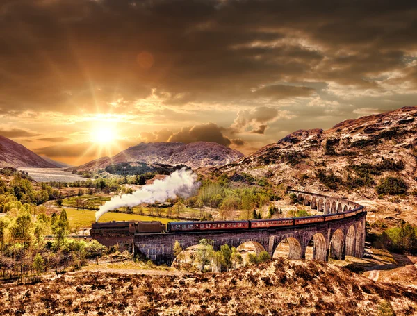 Glenfinnan Viaduto ferroviário na Escócia com o trem a vapor jacobita contra o pôr do sol sobre o lago — Fotografia de Stock