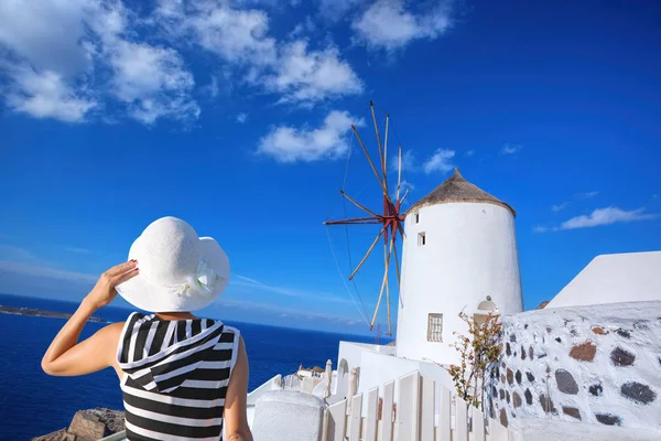 Mujer sosteniendo un sombrero contra el molino de viento en el pueblo de Oia en la isla de Santorini, Grecia — Foto de Stock