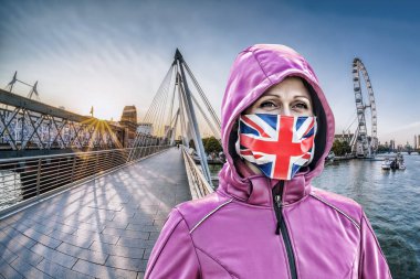 İngiltere 'nin başkenti Londra' da gün batımına karşı Golden Jubilee Köprüsü 'nde İngiliz bayrağına karşı koruma maskesi takan kadın.