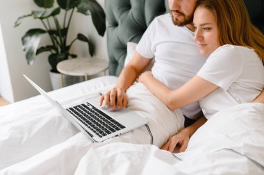 Genç çift yatakta dizüstü bilgisayar kullanıyor. 
