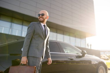 Güneş gözlüklü yakışıklı bir işadamının portresi modern binanın önünde arabanın yanında duruyor.