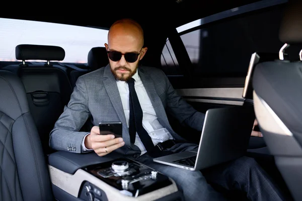 Homem de negócios bonito usando seu telefone celular em um carro moderno com um motorista no centro da cidade. Conceito de negócio, sucesso, viajar, luxo — Fotografia de Stock
