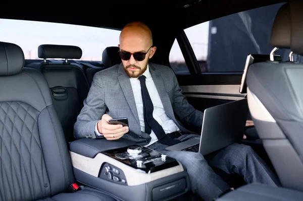 Homem de negócios bonito usando seu telefone celular em um carro moderno com um motorista no centro da cidade. Conceito de negócio, sucesso, viajar, luxo — Fotografia de Stock