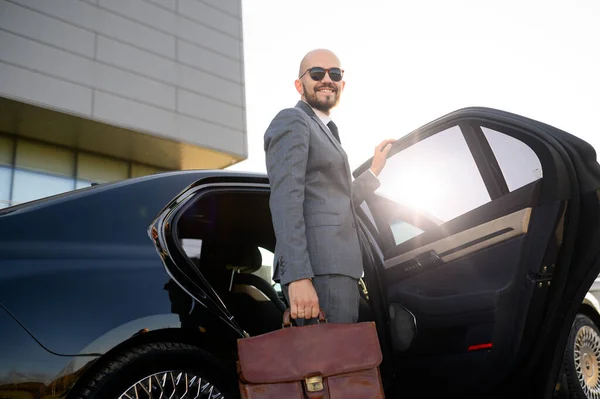 一个戴着太阳镜的英俊商人站在现代建筑立面前的汽车旁边的画像 — 图库照片