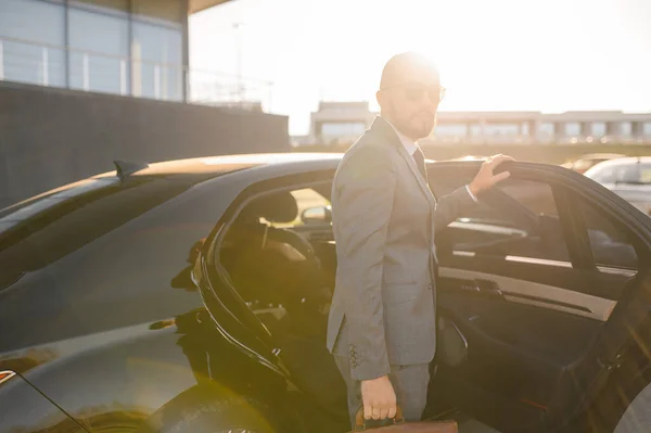 一个戴着太阳镜的英俊商人站在现代建筑立面前的汽车旁边的画像 — 图库照片