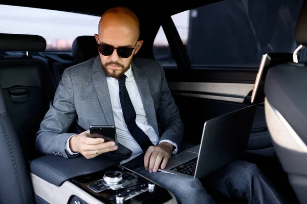 Homem de negócios bonito usando seu telefone celular em um carro moderno com um motorista no centro da cidade. Conceito de negócio, sucesso, viajar, luxo — Fotografia de Stock