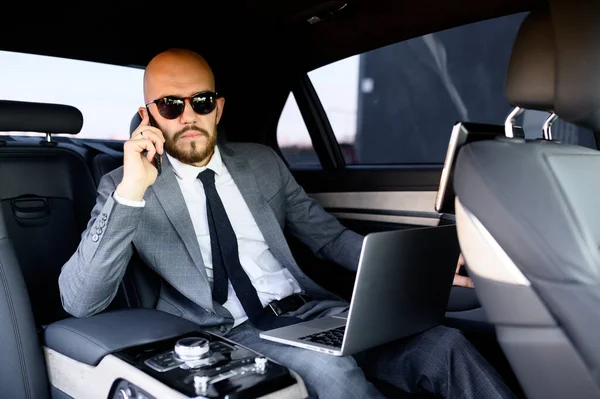 Homem de negócios bonito usando seu telefone celular em um carro moderno com um motorista no centro da cidade. Conceito de negócio, sucesso, viajar, luxo — Fotografia de Stock